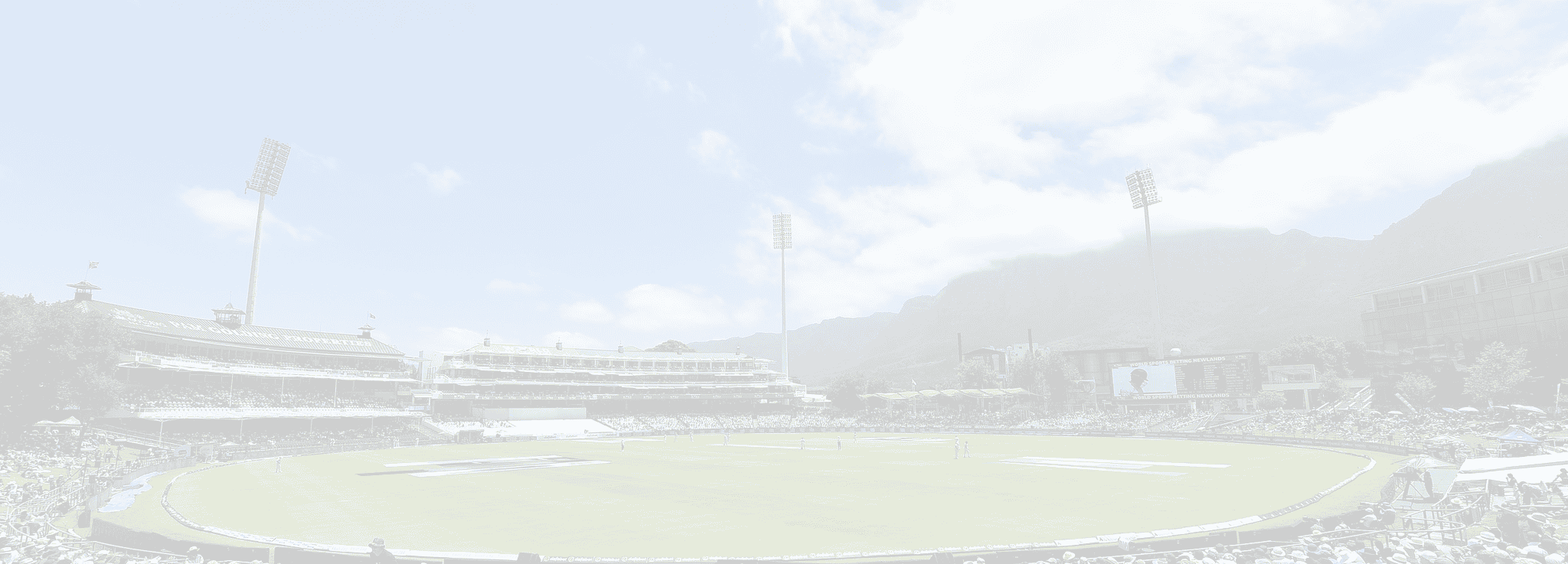 Wide view of a daytime cricket stadium with stands full of spectators, floodlights tall on the field, and mountains in the background